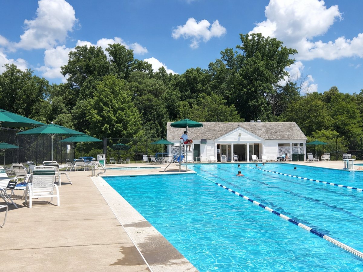 An outdoor lap pool with green umbrellas, lounge chairs and a white bathhouse in front of tall summer woods.