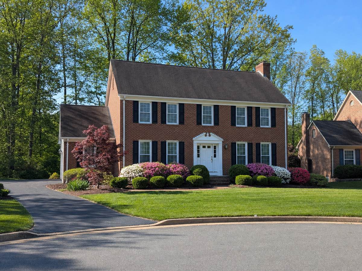 Red-brick center-hall colonial with black shutters, a white pedimented door, pink and white azaleas and a Japanese maple on a cul-de-sac in spring.