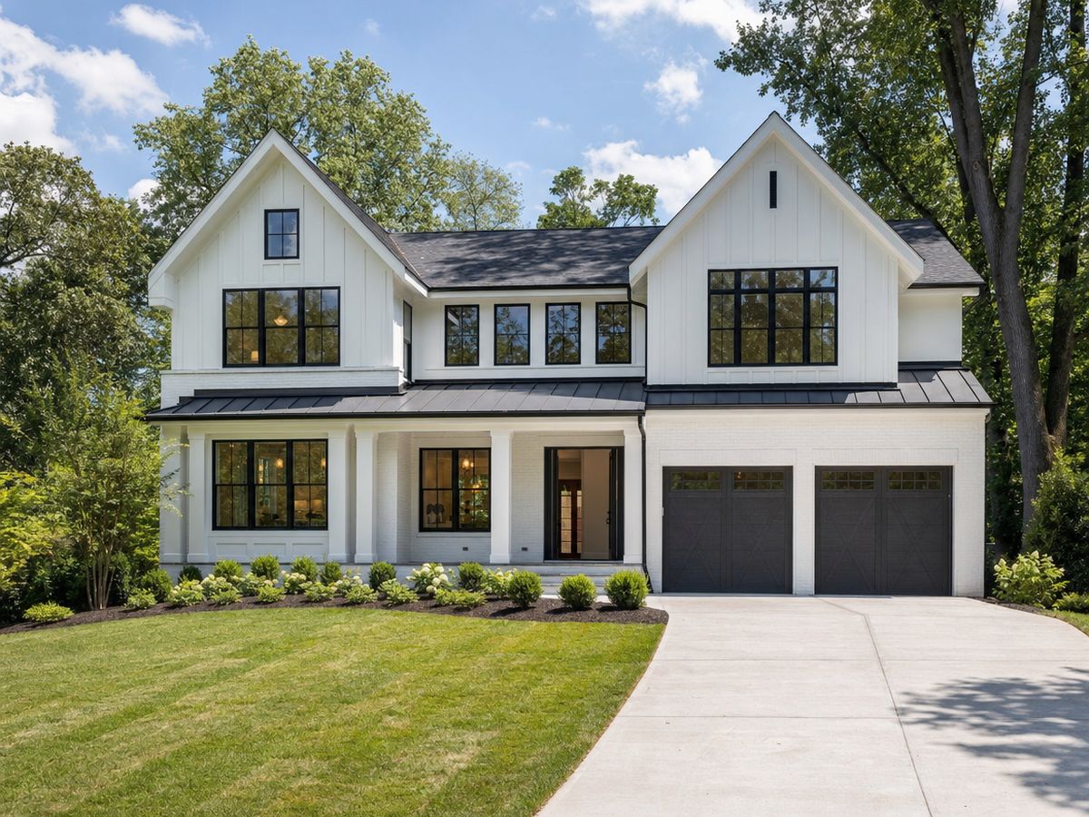 A newly built white two-story house with black-framed windows, a black metal porch roof, an open front door, two dark gray garage doors and a fresh lawn under mature trees.