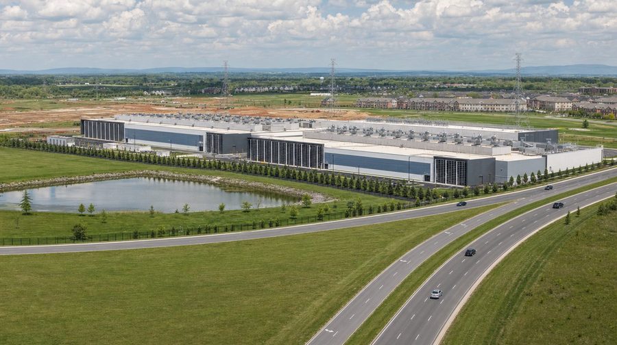 A long data center campus beside a divided parkway, a pond and green fields