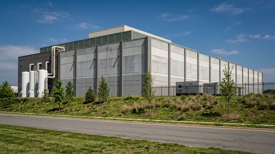A gray data center wall with white tanks beside it, seen from a quiet road