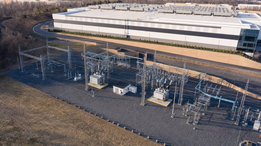 An electric substation with transformers and steel frames beside a long white data center