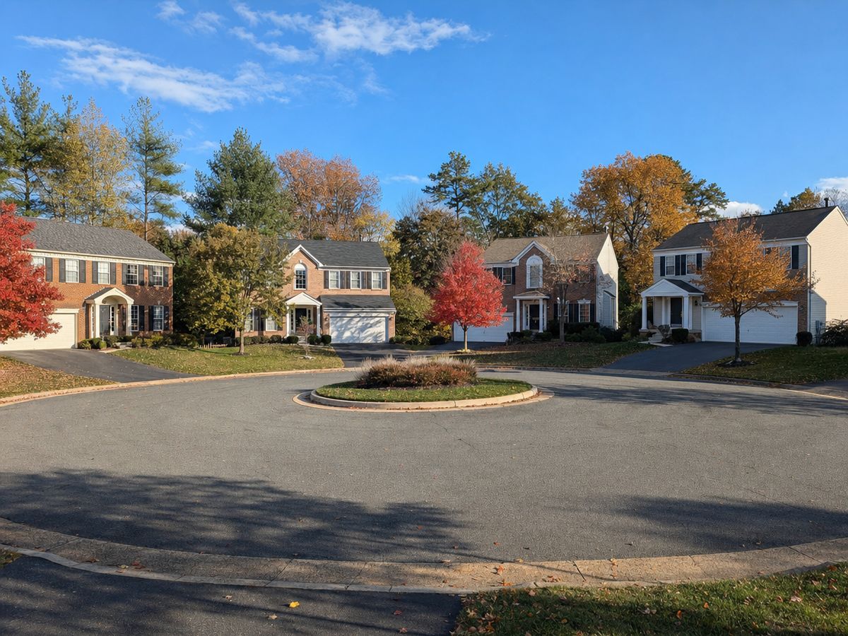 A round cul-de-sac with a small planted island surrounded by brick-front two-story colonials with white garage doors and red and orange autumn trees.