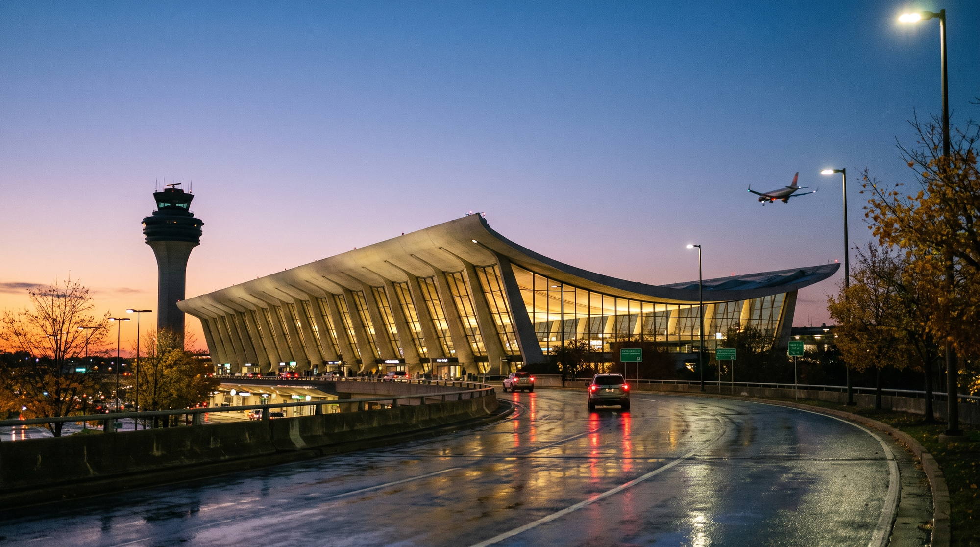 The swooping main terminal and the original control tower of Dulles Airport at dusk, the glass walls glowing under the curved concrete roof, with a jet on approach overhead and car lights on the wet, curving road in front
