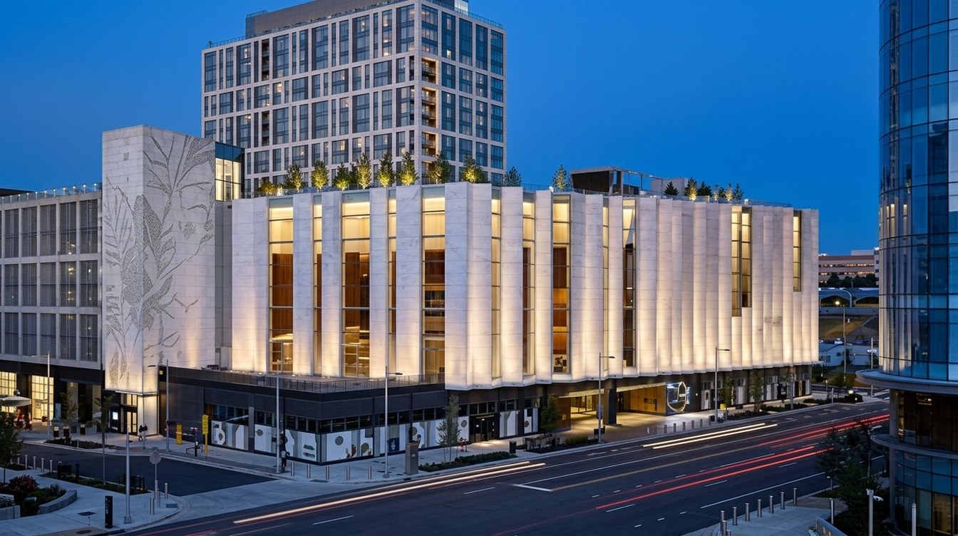 A white pleated performance hall lit warm from below, with a leaf-patterned end wall, rooftop trees, a hotel tower behind and a curved glass tower at the edge, as car lights streak past at dusk