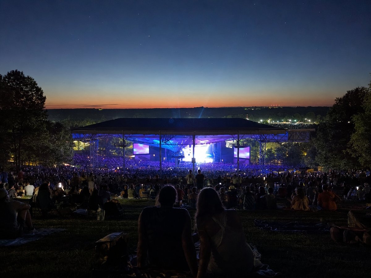 A large outdoor concert crowd on a grass lawn at dusk, looking down at a glowing roofed stage pavilion under a deep blue sky.