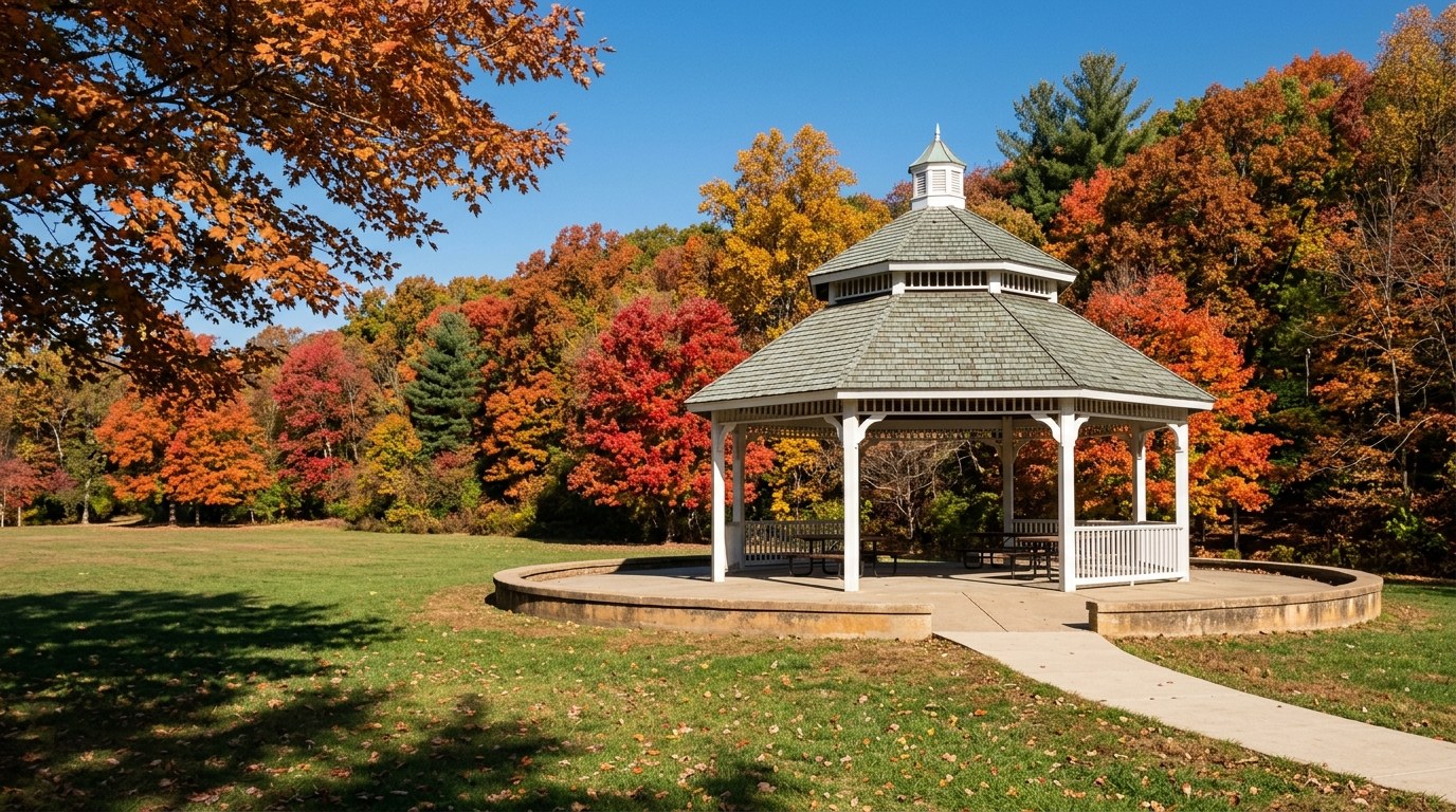 A white octagonal gazebo with a green-gray two-tier roof and a small cupola on a sunny lawn, backed by red, orange and gold fall woods.